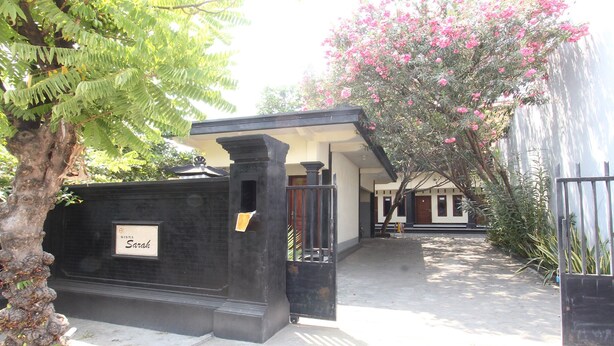 A black gate with a nameplate reading "Sarah," a pathway leading to a building, and a tree with pink flowers. - OYO Life 93155 Wisma Sarah (Tanjung)
