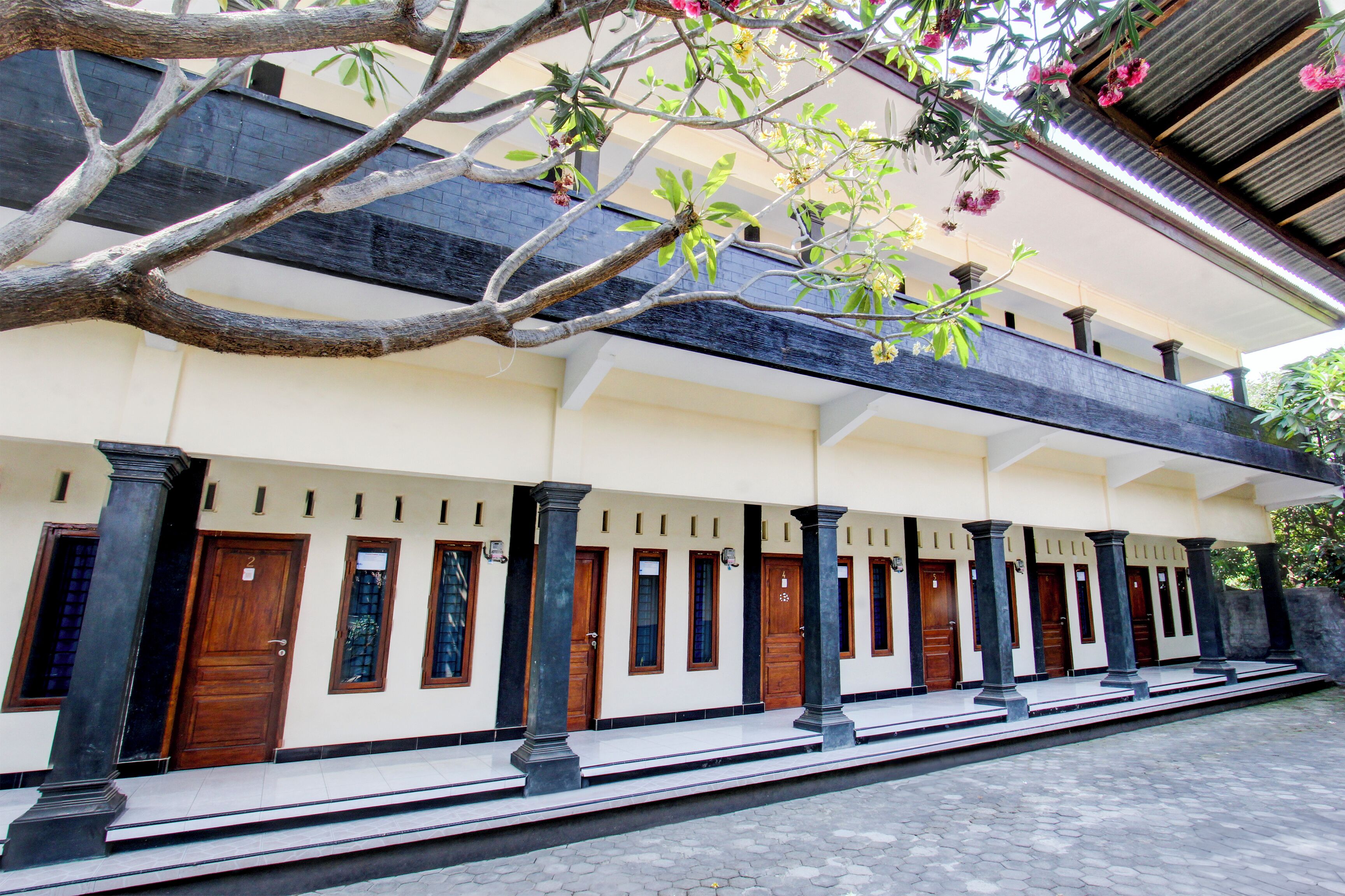 Un edificio con varias puertas y columnas, un área de entrada con baldosas y un árbol con flores.