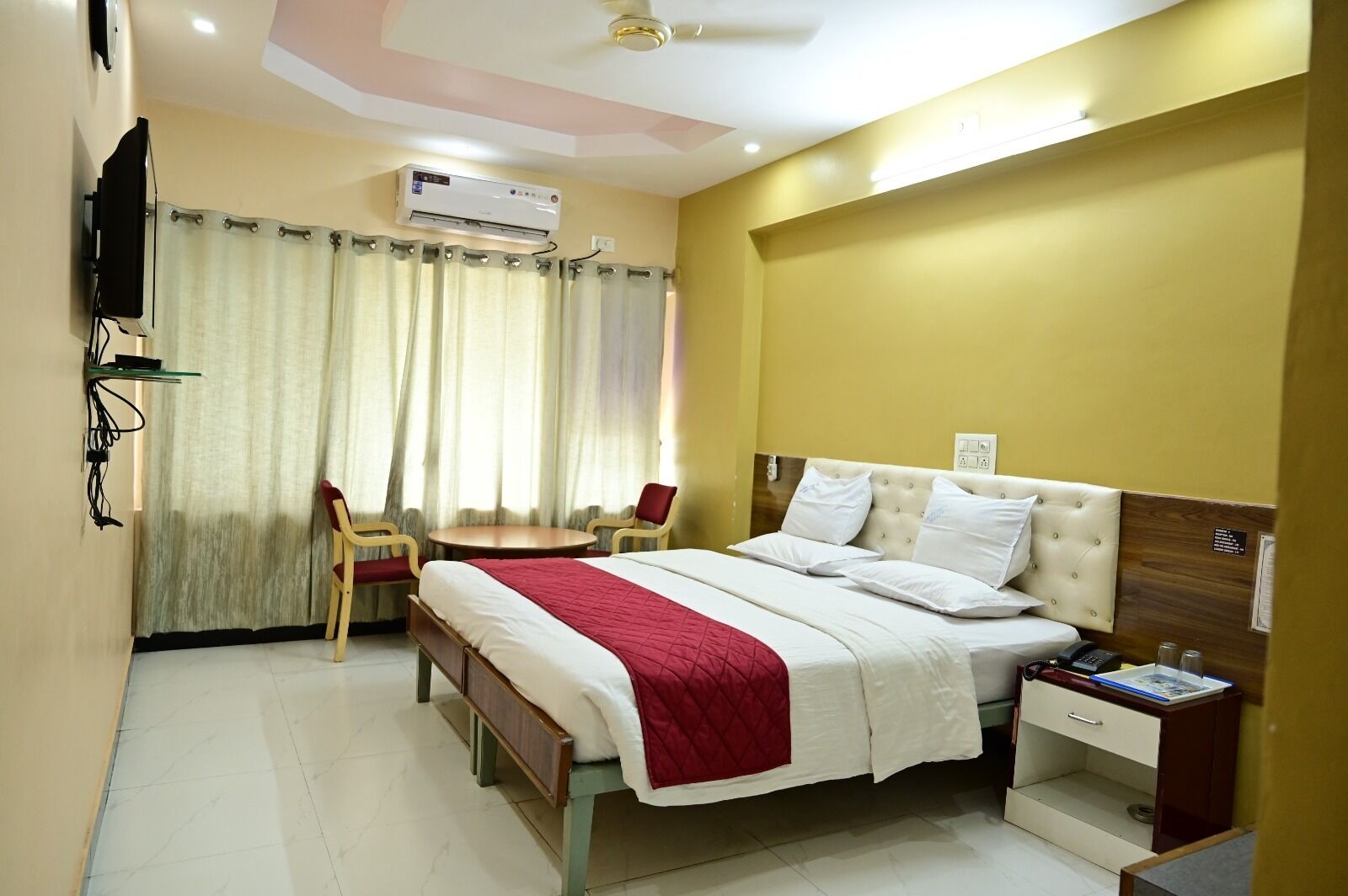 A hotel room with a bed, bedside table, chair, and an air conditioning unit.