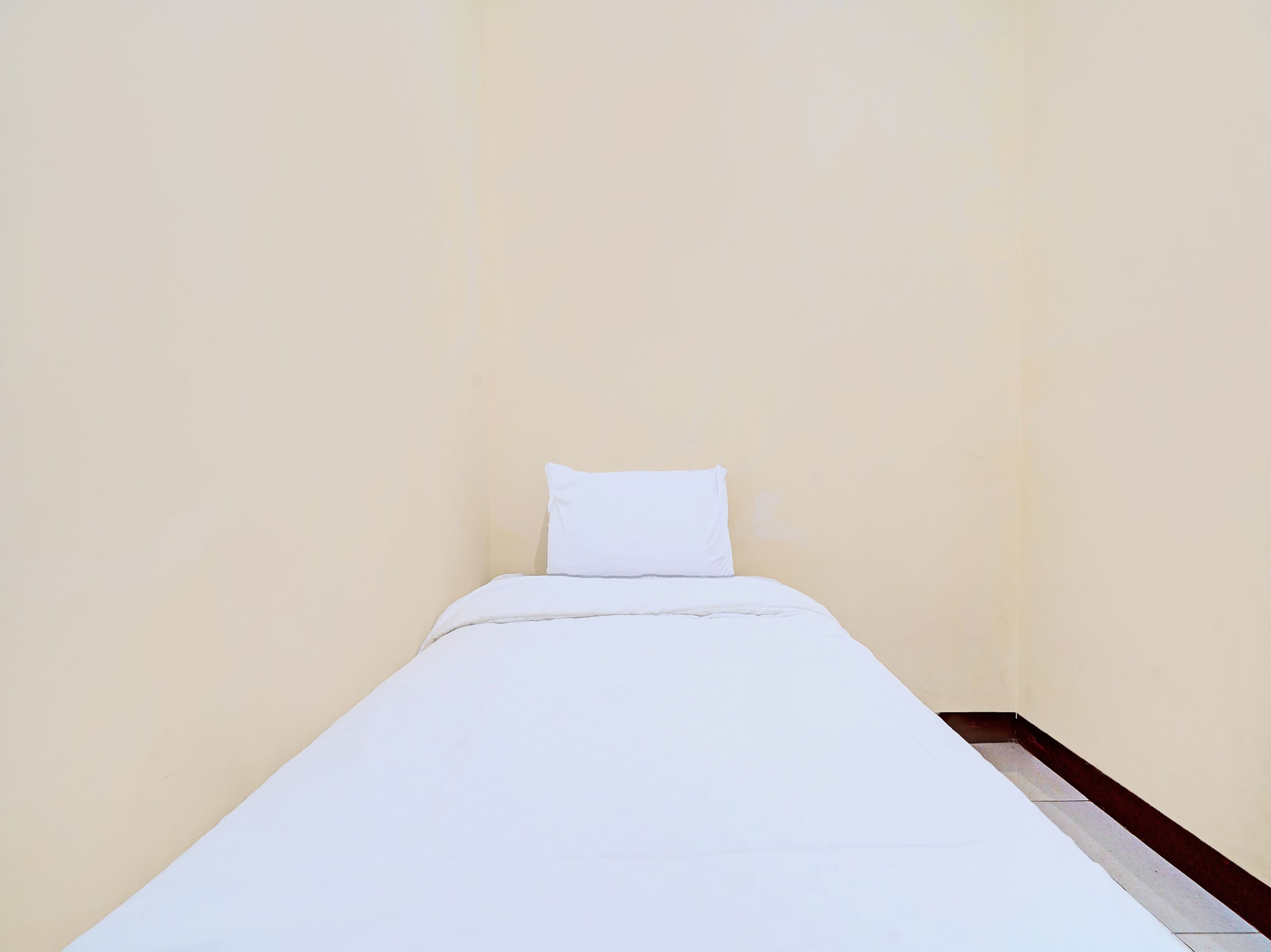 A small, single-bed room with a plain white bed and a white pillow. The walls are a light beige color, and the floor appears to be tiled.