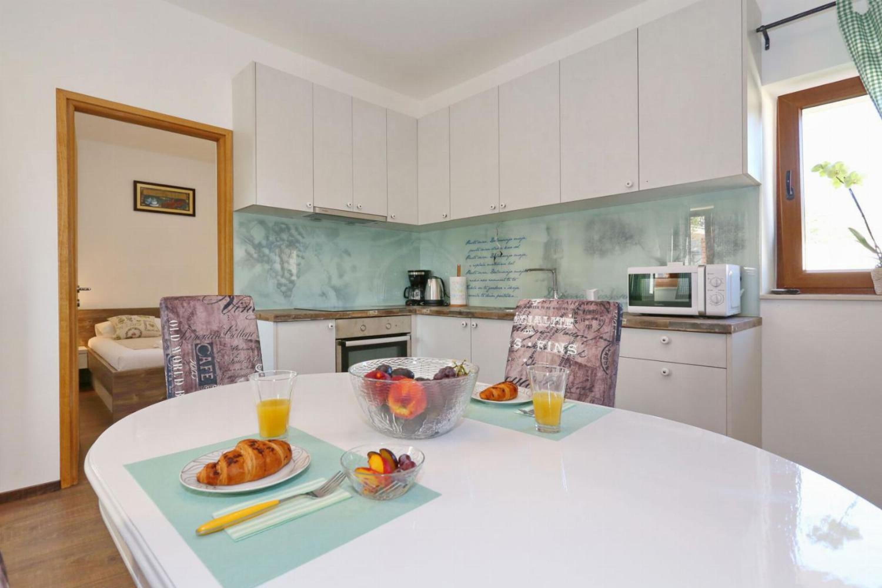 A modern kitchen with white cabinets, a microwave, and a coffee maker. There is a dining table with a fruit bowl, croissants, and juice glasses.