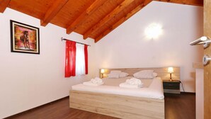 Un dormitorio con cama de madera, ropa de cama blanca, cortina roja y blanca, un cuadro en la pared y una mesita de noche con lámpara.