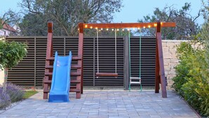 Un parque infantil con tobogán, columpios y un banco bajo una pérgola de madera con luces colgantes.