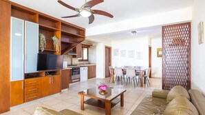 A modern living room with a sofa, coffee table, and TV. There is a kitchen area with cabinets and a dining table. - Villa Sunlove in Peyia (Pegeia)