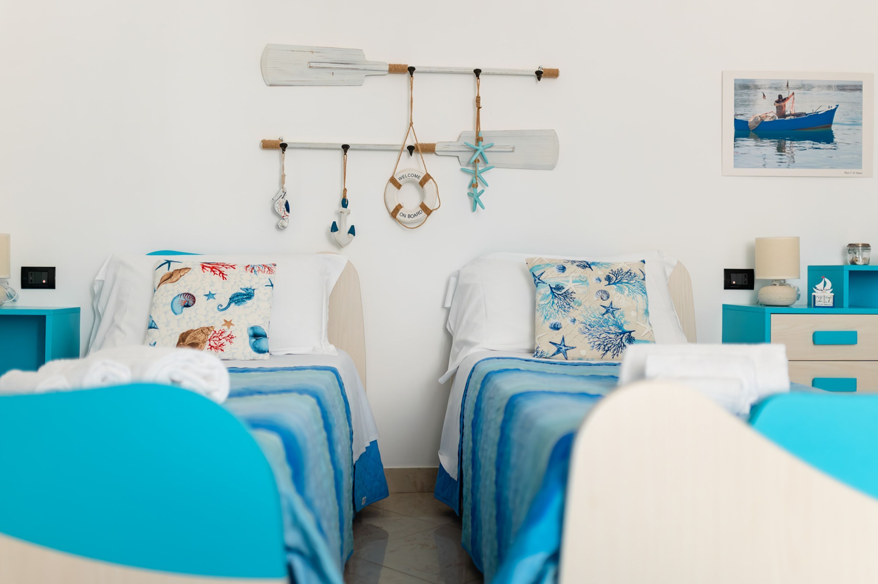A room with two beds, a wall-mounted life preserver, a paddleboard, and a framed picture of a boat.