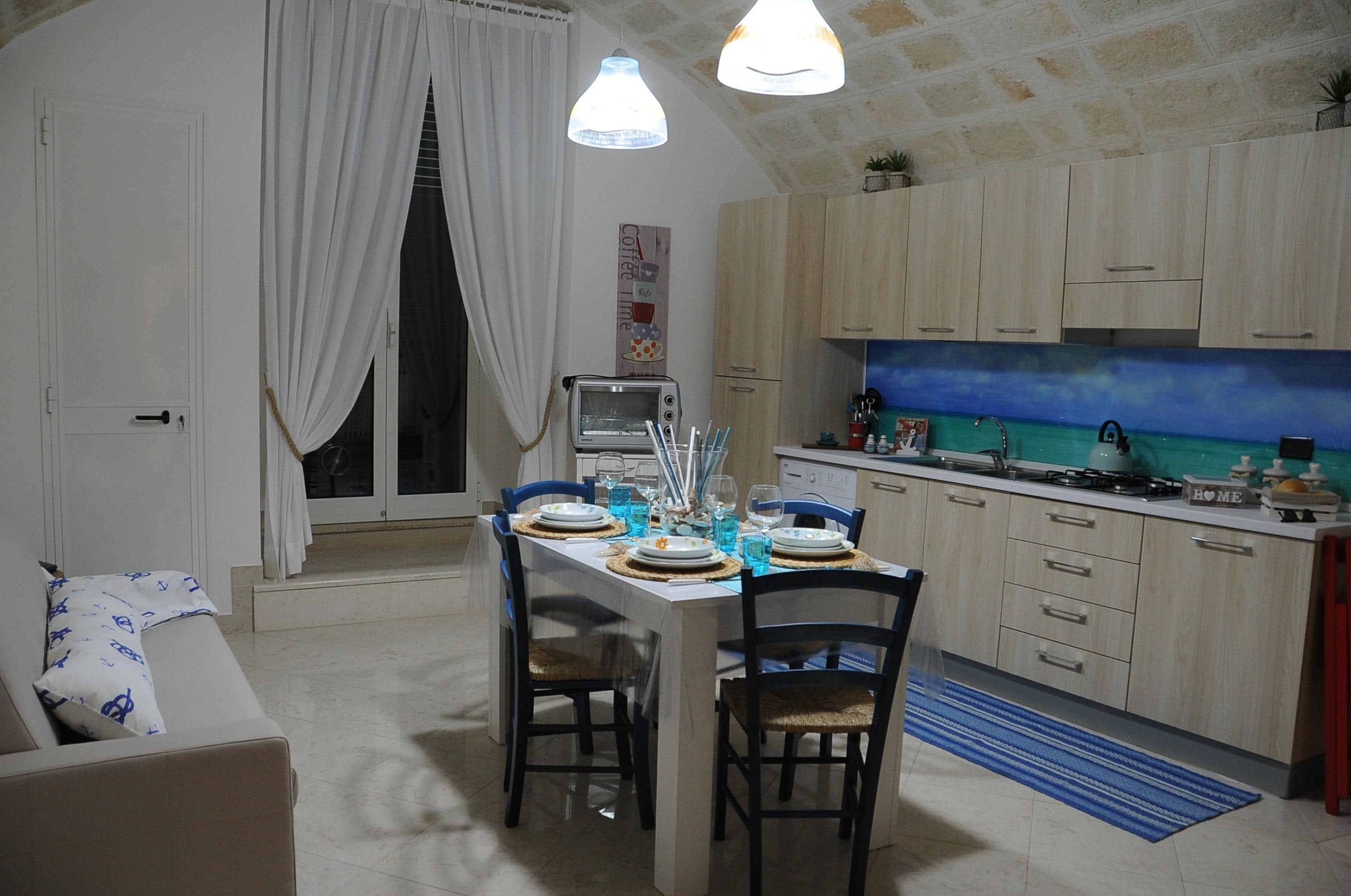 Cuina compacta amb zona de dinar, forn encastrat i una finestra petita amb cortines.