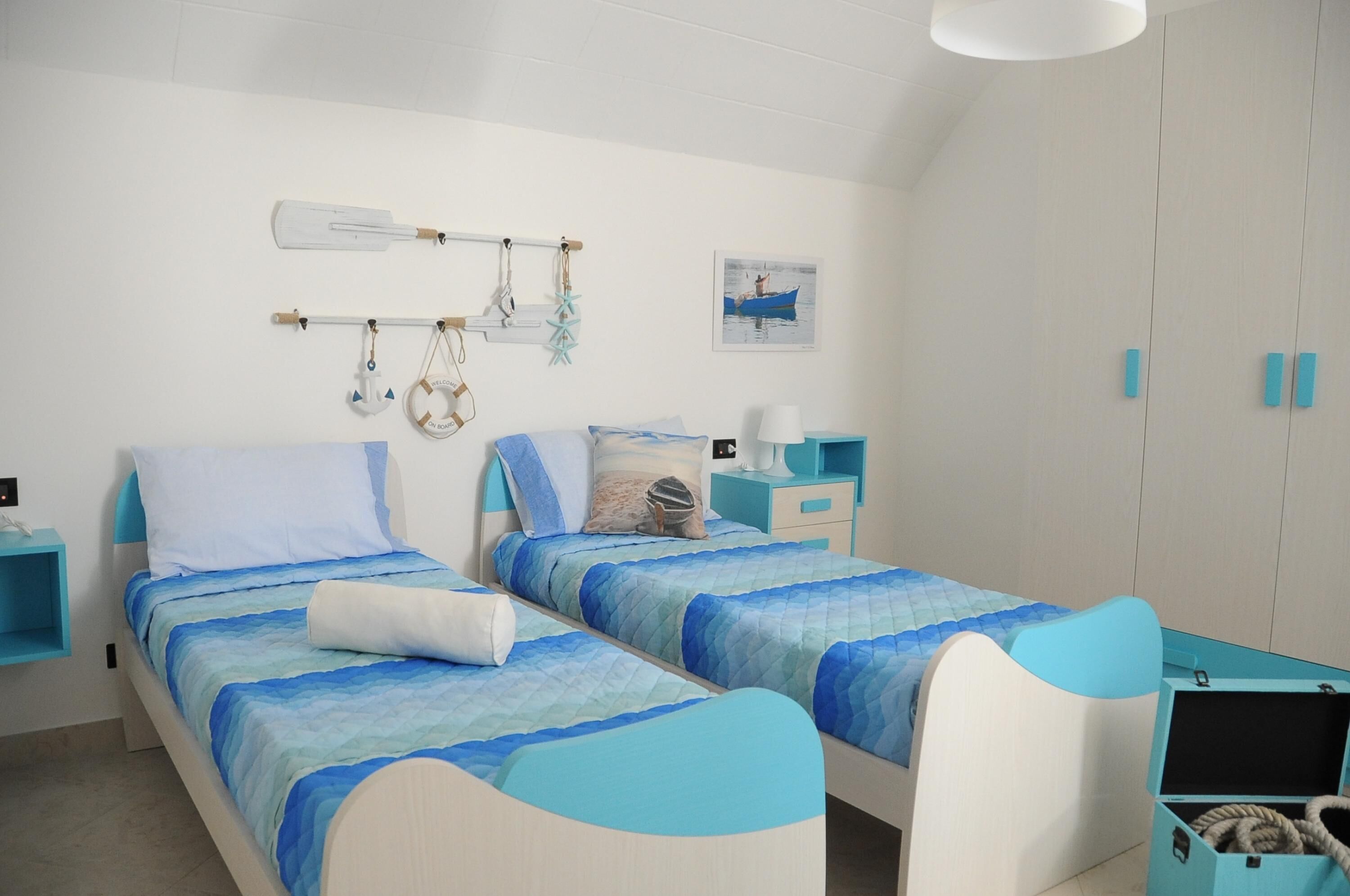 Un dormitorio con dos camas, un perchero con objetos, un cuadro enmarcado y un pequeño escritorio con una lámpara.