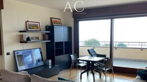 Een moderne woonkamer met een bank, een eettafel met stoelen, een televisie en een groot raam met uitzicht op de omgeving.