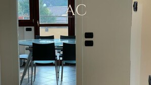 Uitzicht door een deur op een eetruimte met een glazen tafel en stoelen, een betegelde vloer en een raam met zicht op gebouwen en bomen.