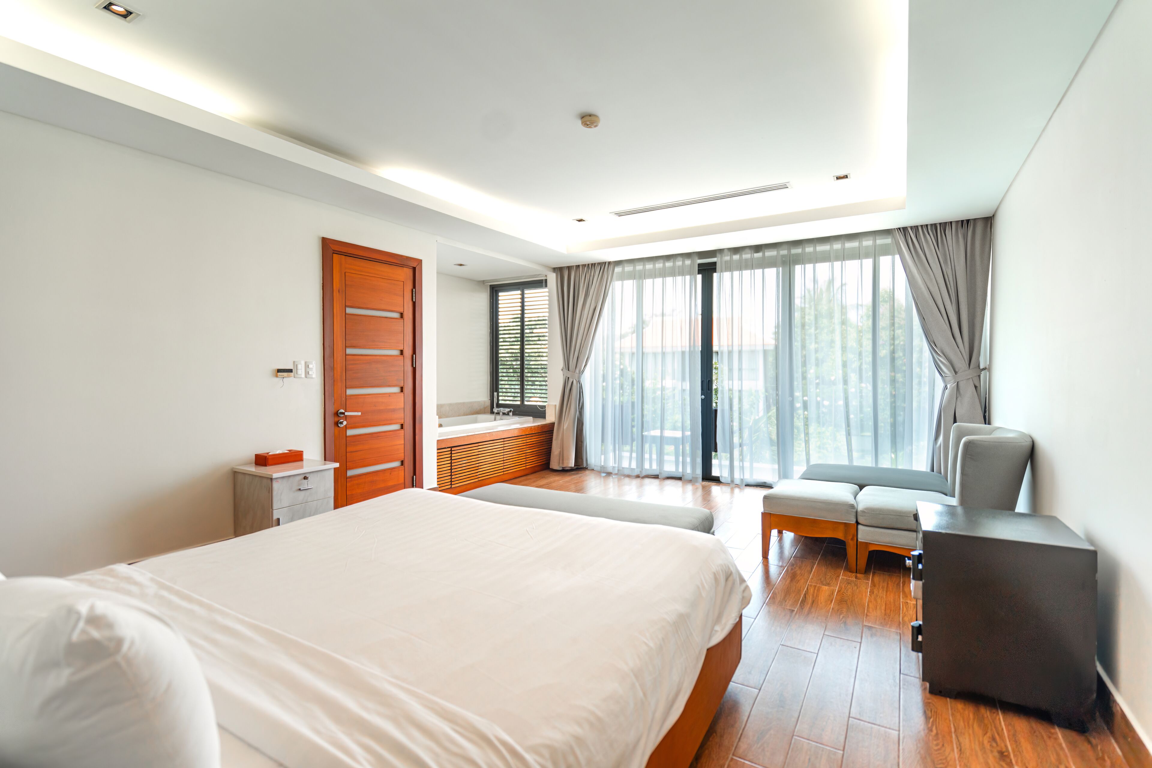 A modern hotel room with a large bed, a seating area with a sofa and armchairs, a small table, and a large window with curtains.