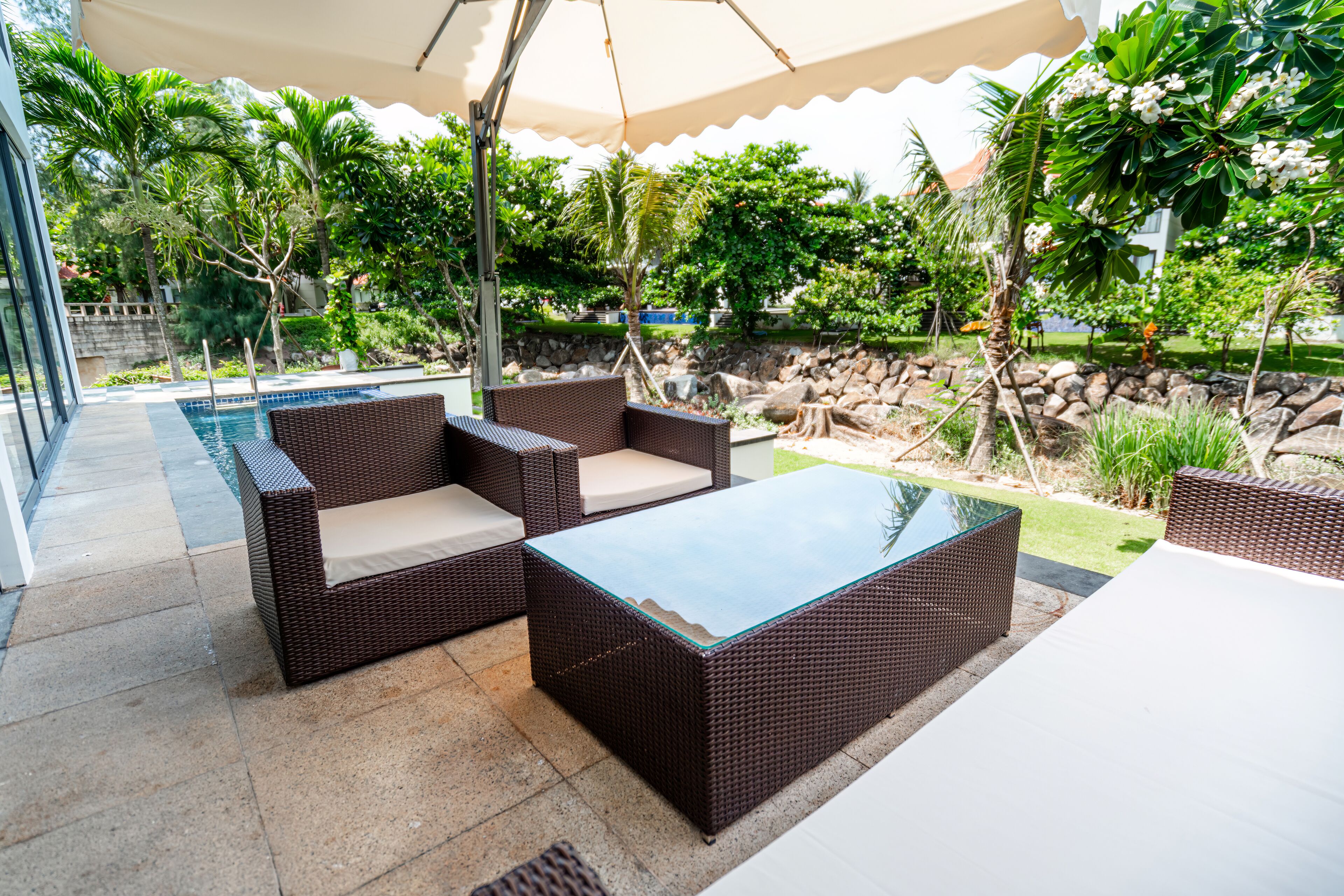 An outdoor patio with wicker furniture, a glass-top table, and a pool in the background.