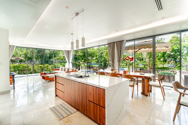 A modern kitchen with a large island, wooden cabinets, and a view of a lush garden through large windows.