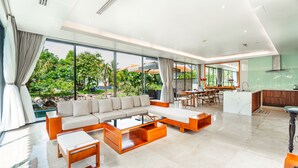 A spacious living area with a large sofa, coffee table, and dining table. A kitchen is visible in the background with a view of greenery outside.