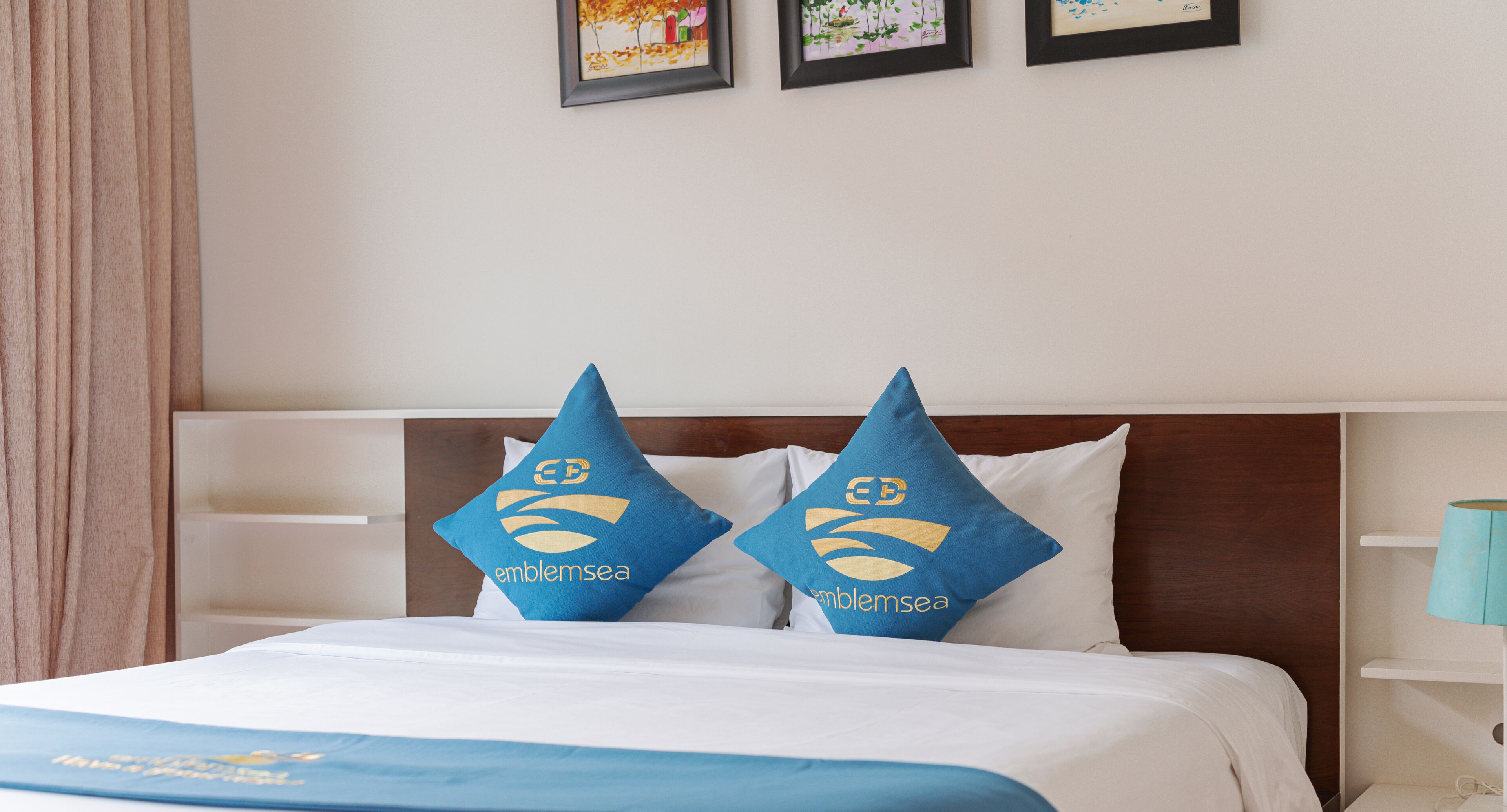 A hotel room with a neatly made bed, two pillows with a logo, a wooden headboard, and a nightstand with a lamp.