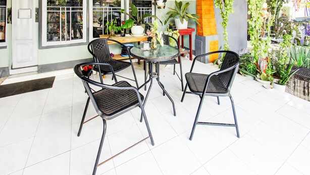 Un área de patio con una mesa redonda y cuatro sillas, rodeada de plantas en macetas y un piso de baldosas.