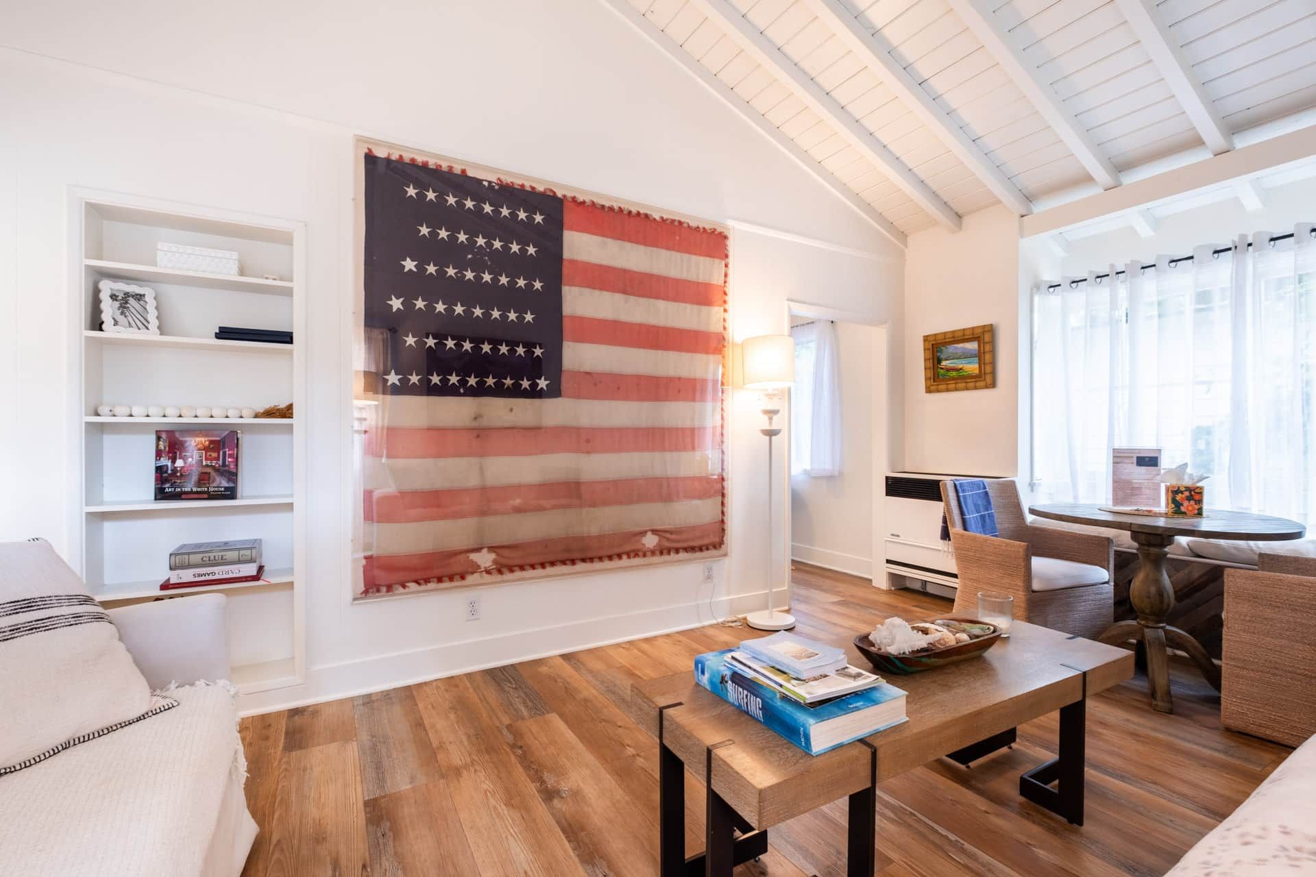 Una habitación con una gran bandera estadounidense, piso de madera, una mesa de comedor con libros, una silla de mimbre y una estantería.