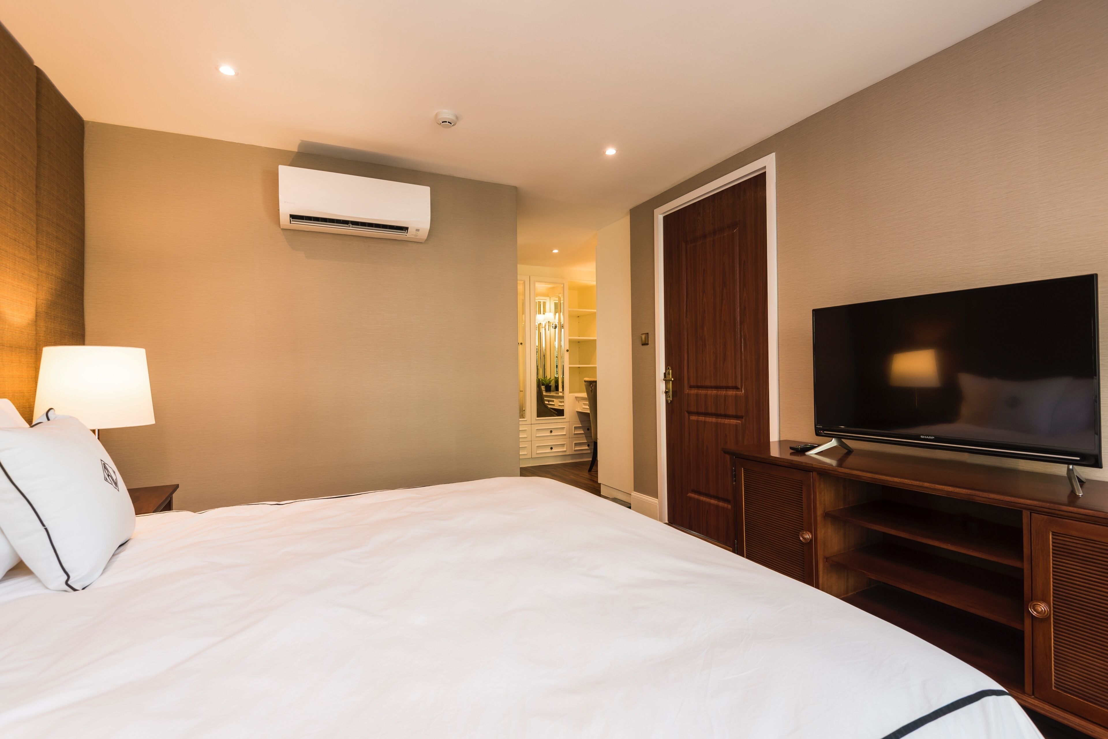 Quarto de hotel com uma cama, televisão sobre um suporte de madeira, aparelho de ar condicionado na parede e porta que dá para o banheiro.