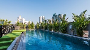 Uma piscina com superfície de água azul cristalina, rodeada por vegetação bem cuidada e edifícios modernos ao fundo.
