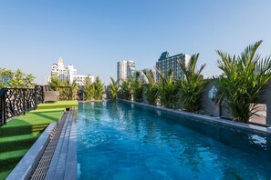 A swimming pool with a clear blue water surface, surrounded by well-maintained greenery and modern buildings in the background.
