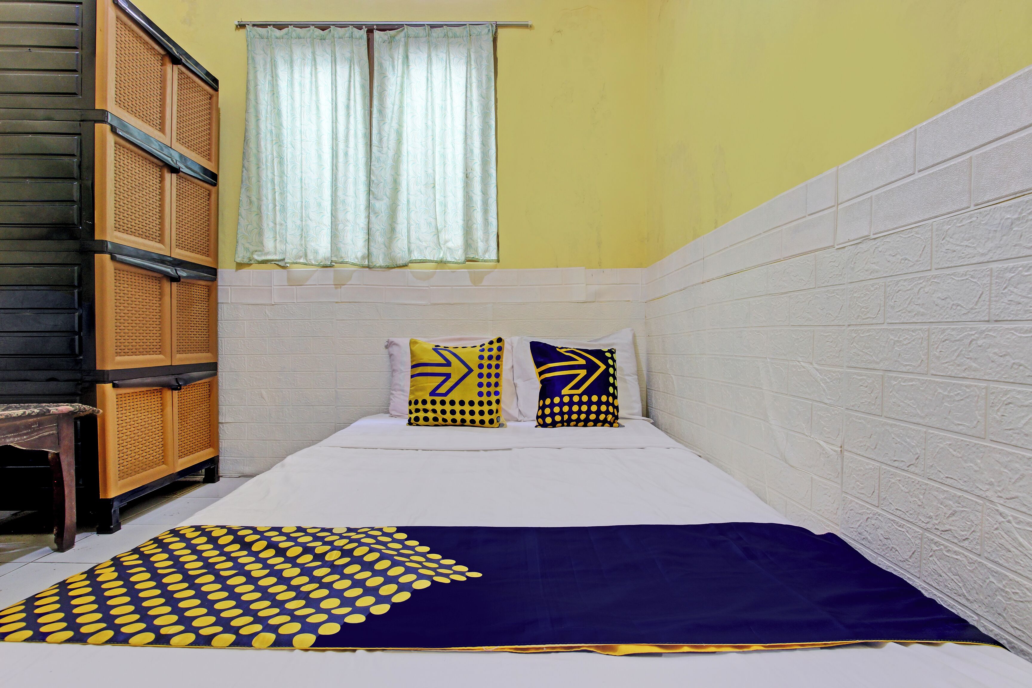 A small, narrow room with a bed, two pillows, a patterned blanket, a window with curtains, and a wall with a textured pattern.