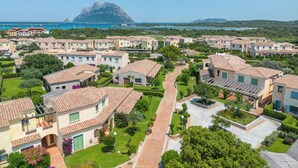 A residential area with terracotta-roofed houses, a central paved walkway, and a backdrop of a mountainous landscape and a body of water. - I Fari Apartments 600m From The Beach (Loiri Porto San Paolo)
