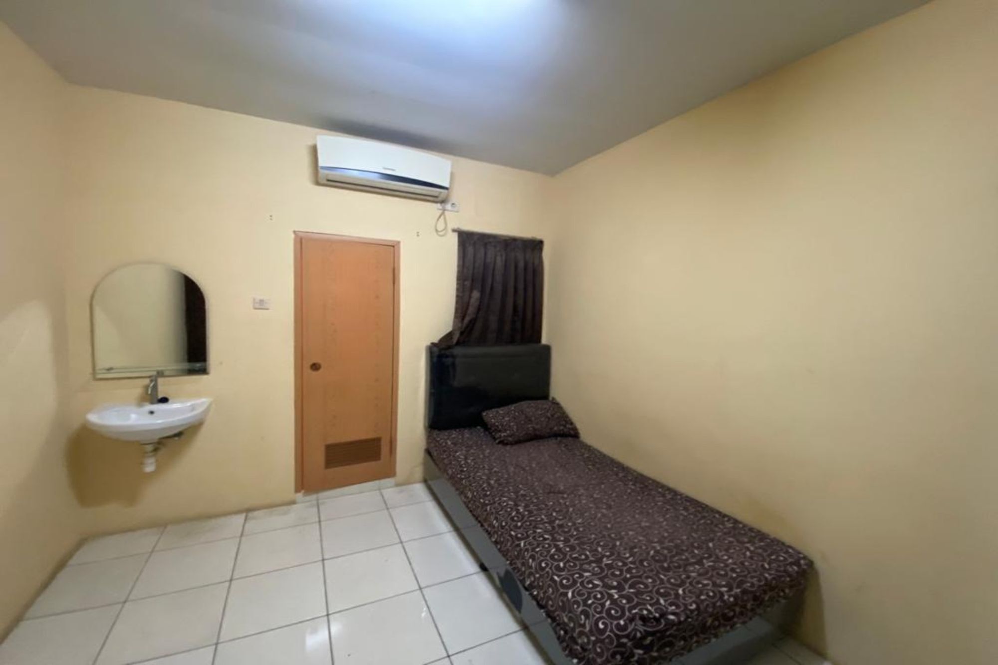 A small, single-room accommodation with a bed, a bedside table, a mirror, a sink, and an air conditioning unit.