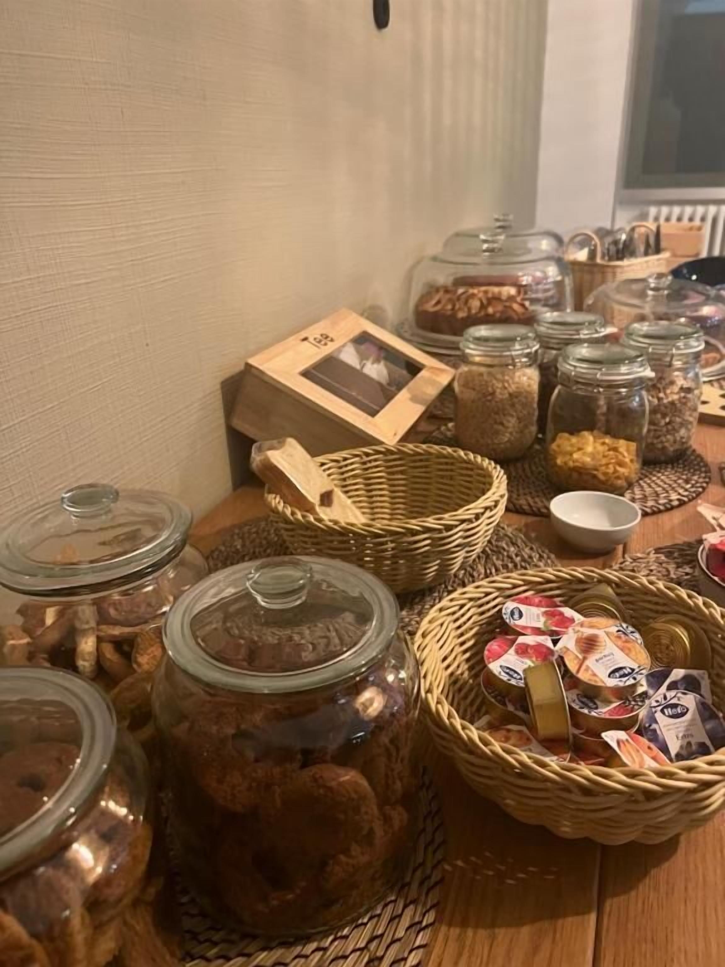 Et bord med forskellig slags mad og beholdere, herunder glas med nødder og tørret frugt, kurve med snacks, og et indrammet foto.