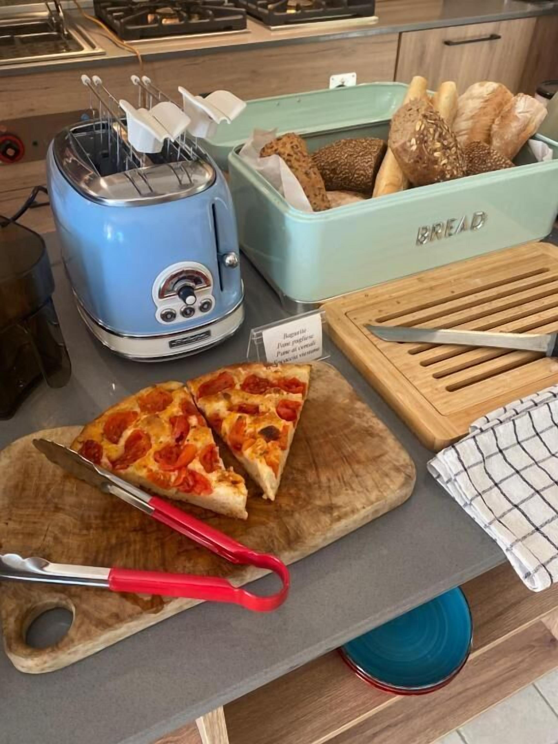 A kitchen counter with a blue toaster, a bread box, a wooden cutting board with pizza slices, and a pair of red-handled tongs.