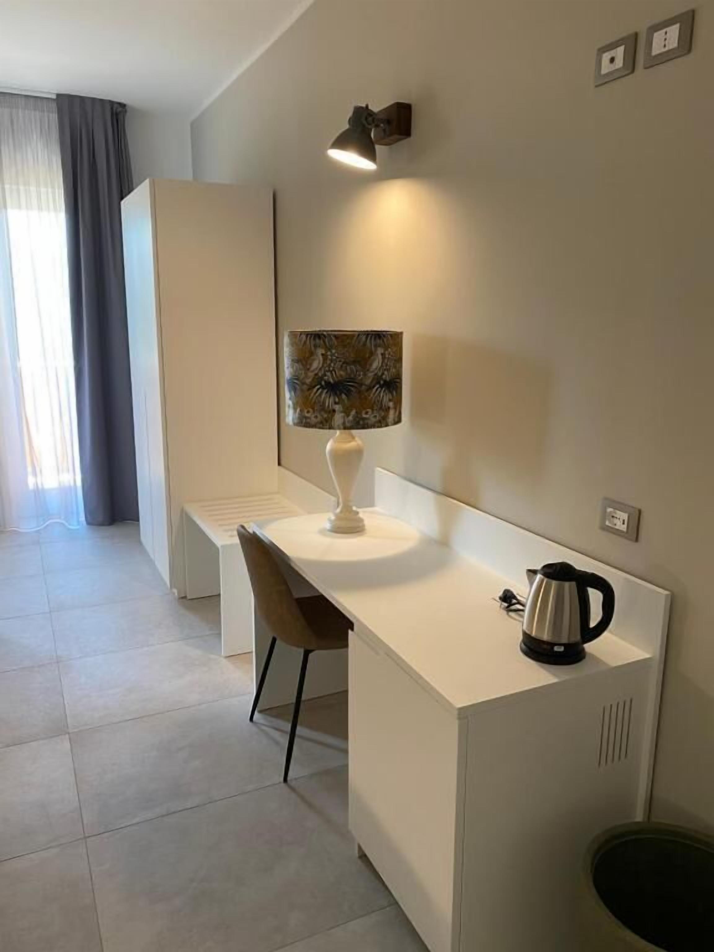 A modern, minimalist hotel room with a white desk, a chair, a coffee maker, and a painting on the wall.