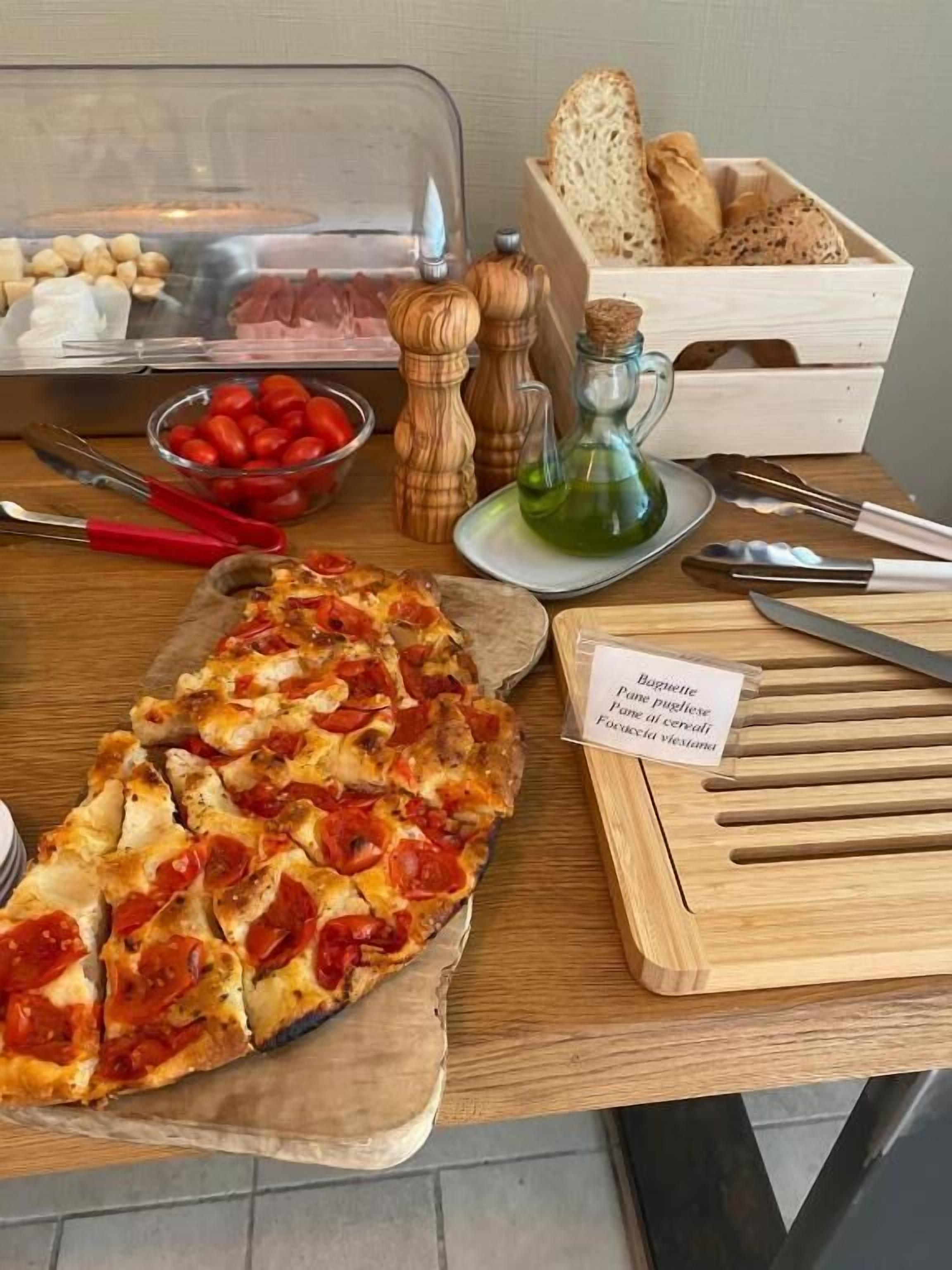 A wooden table with a pizza, a bowl of cherry tomatoes, a pepper mill, a bottle of olive oil, and a basket of bread.