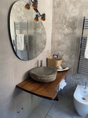 A bathroom with a round mirror, a wooden vanity, a stone sink, a white pedestal sink, and a towel rack. - La Corte del Sole (Vieste)