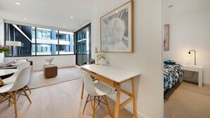 Una sala moderna con un escritorio blanco, un sofá y una cama con colcha de flores azules y blancas. Hay un ventanal con vista a un edificio y una obra de arte enmarcada en la pared.