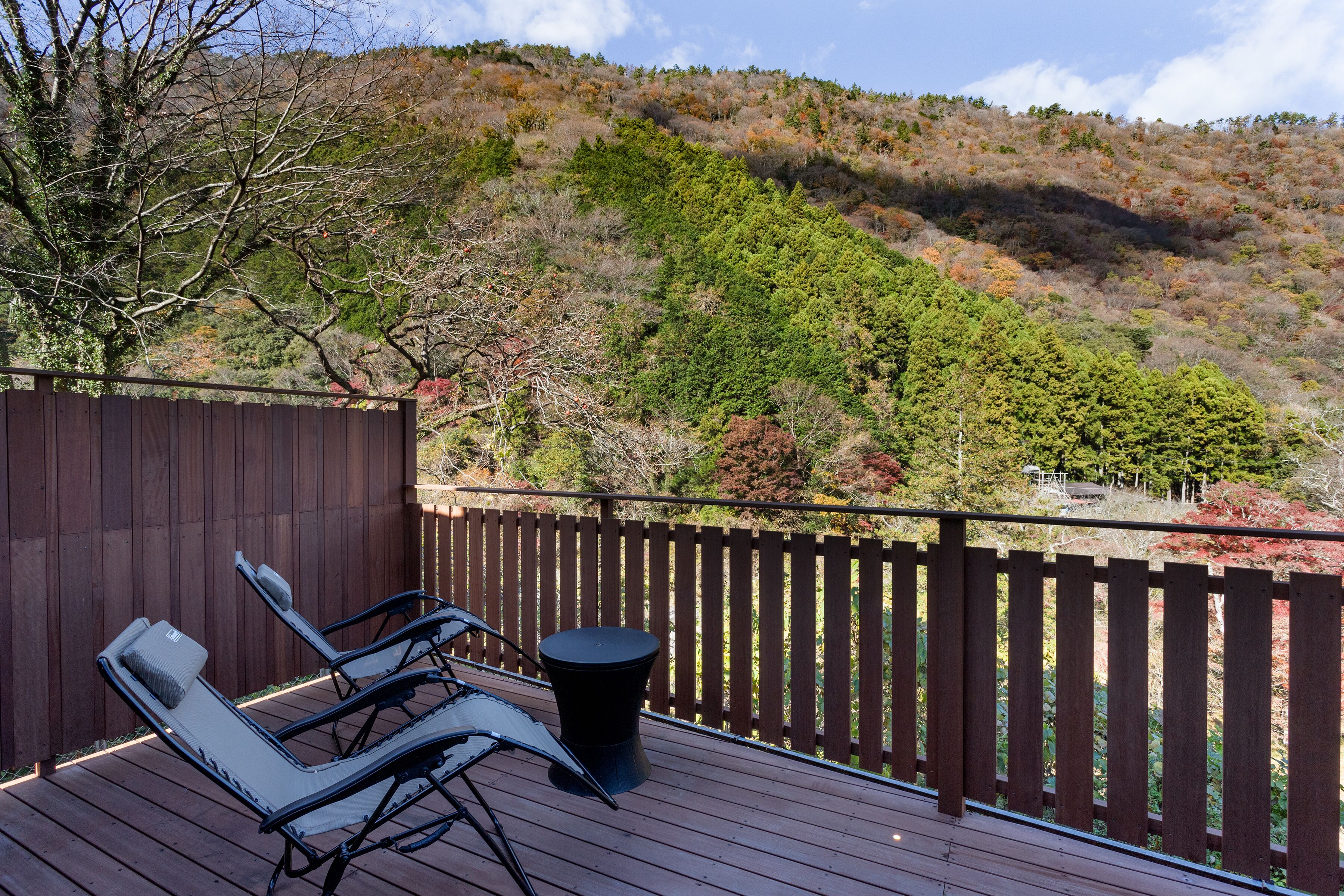 A deck with two lounge chairs and a small table, overlooking a hilly landscape with trees and a wooden fence.