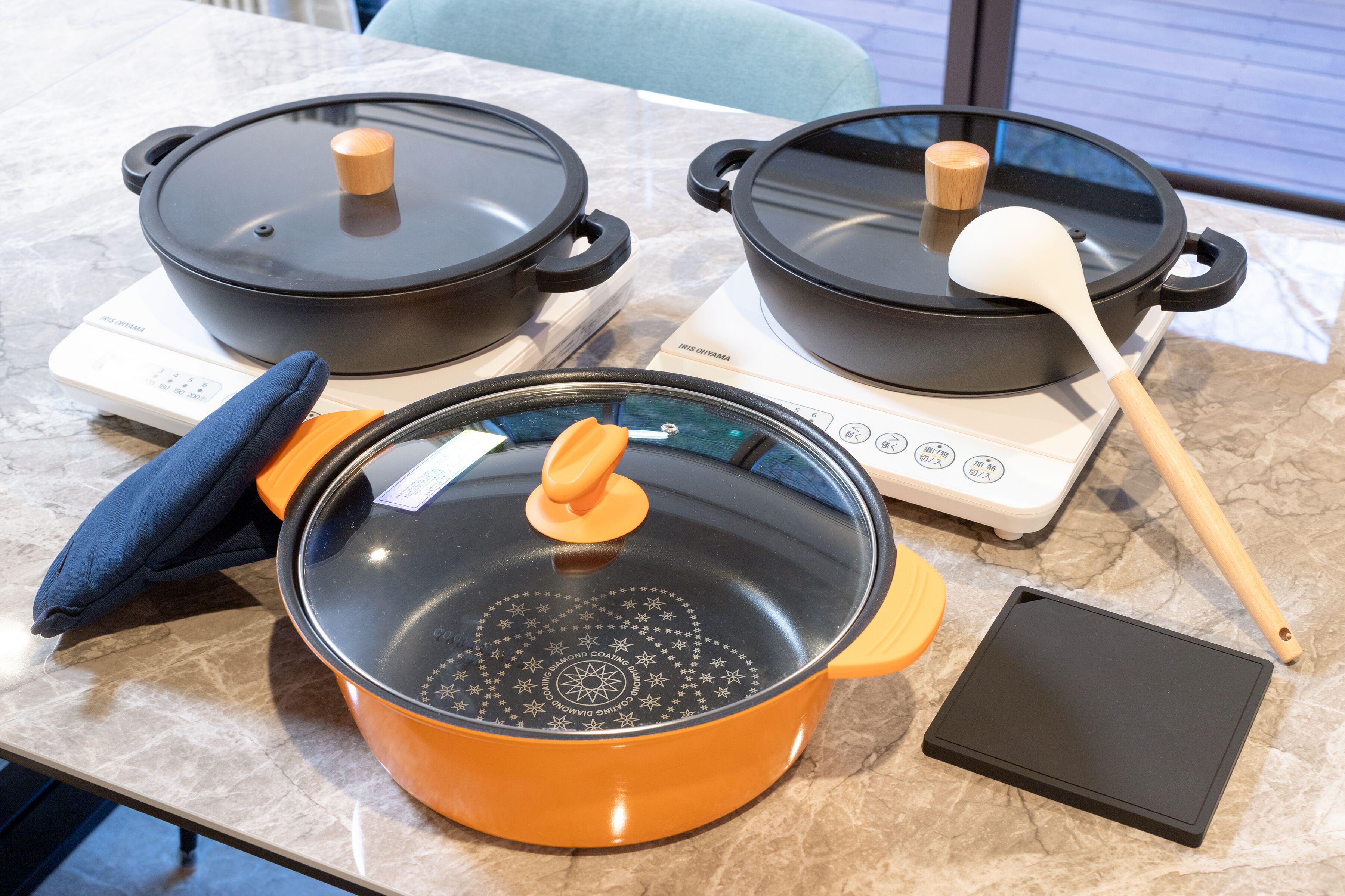 Duas cozinhas elétricas com tampas, uma espátula, uma panela com tampa e um tabuleiro preto numa bancada de cozinha.