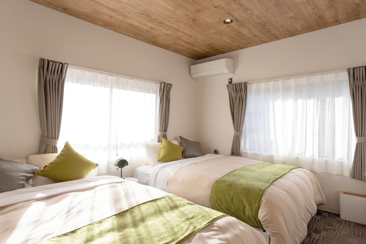 A hotel room with two beds, a wooden ceiling, an air conditioning unit, and large windows with curtains.