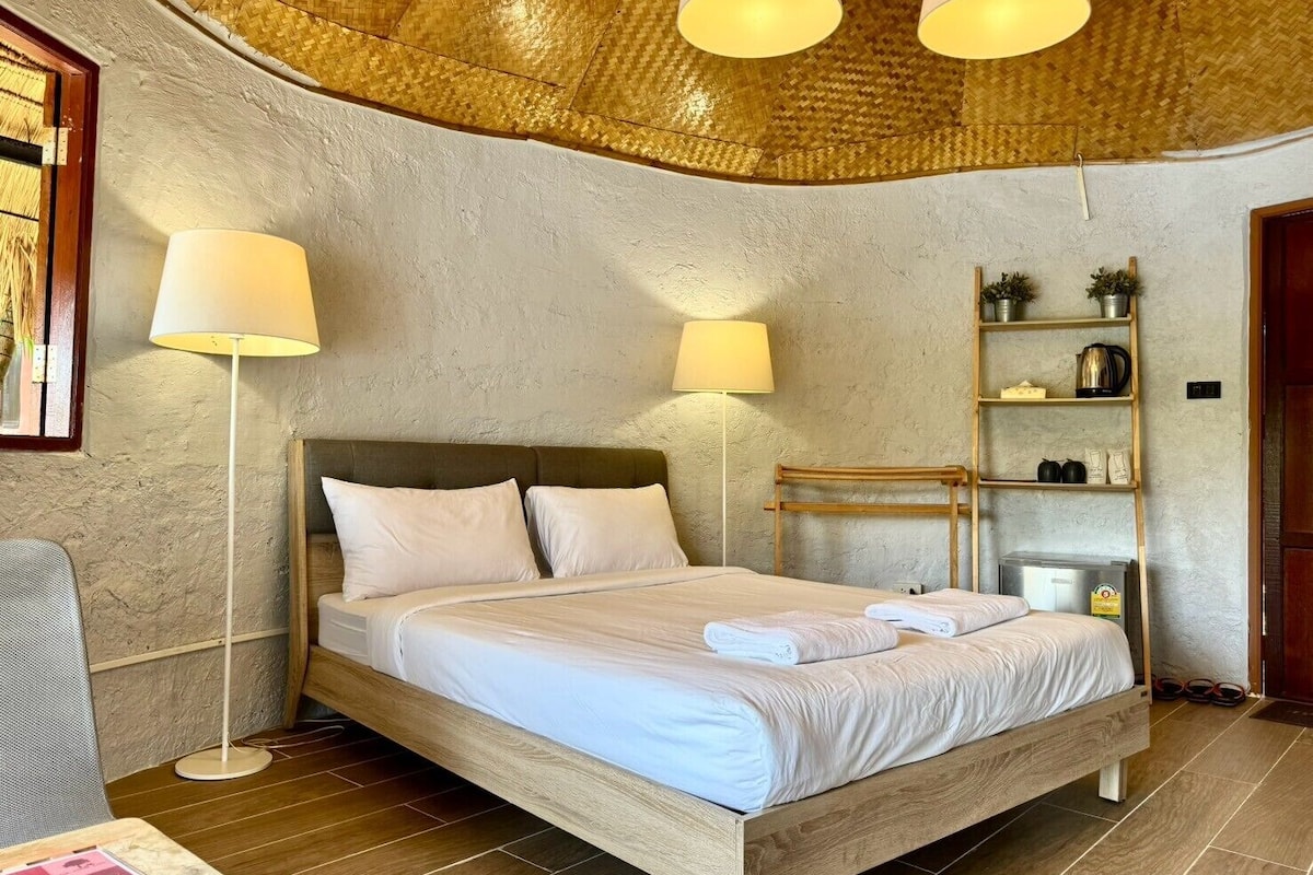 Un dormitorio con cama de madera, ropa de cama blanca, dos lámparas de noche, estante con artículos de aseo y techo tejido.