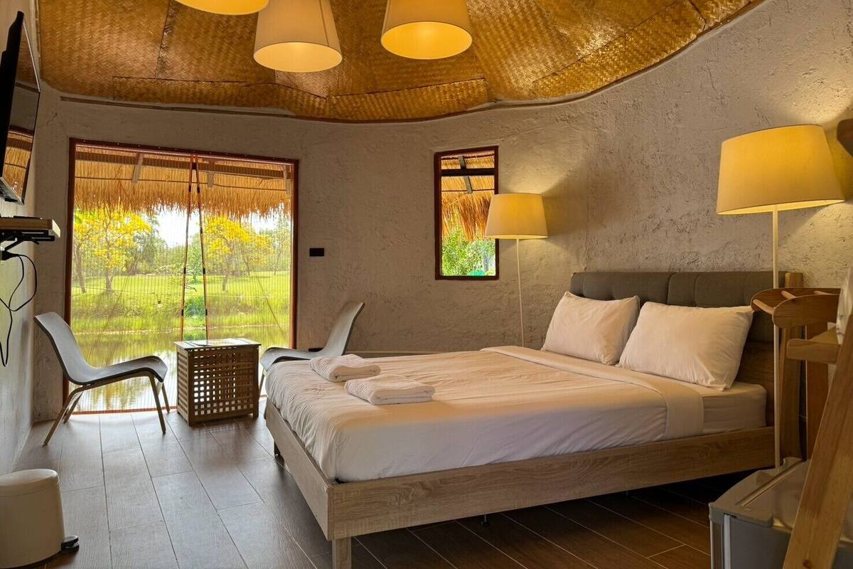Un dormitorio con cama, dos lámparas de noche, una silla, una mesita y ventana con vistas a zonas verdes.