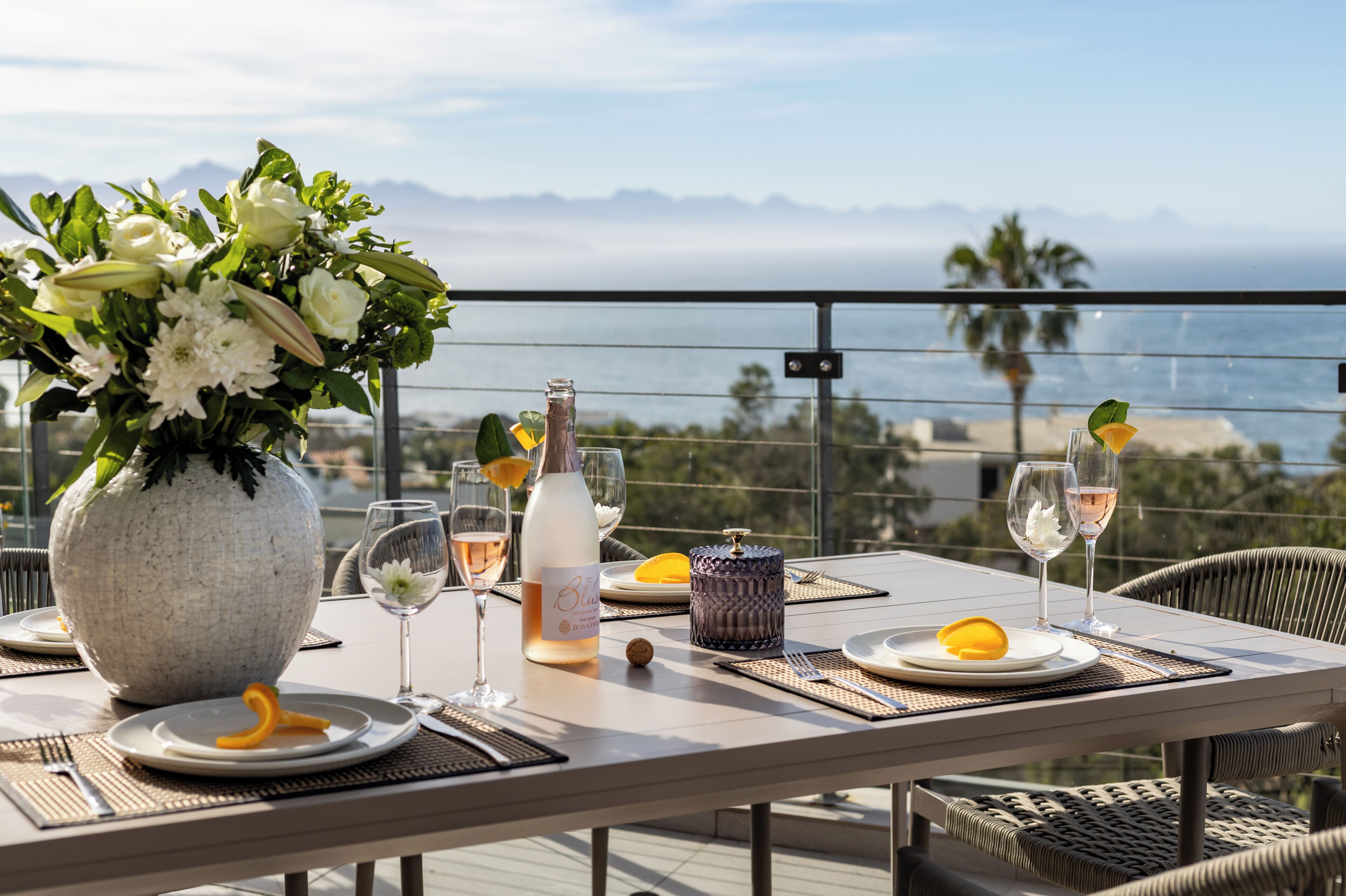 A table set for dining with a view of the ocean, a bottle of wine, and a vase of flowers.