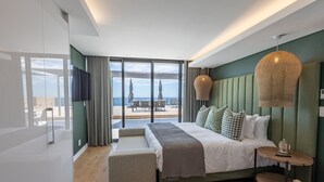 A modern hotel room with a large bed, a small table, a bench, and a view of the sea through large windows. - Plett Quarter Apartments (Plettenberg Bay)