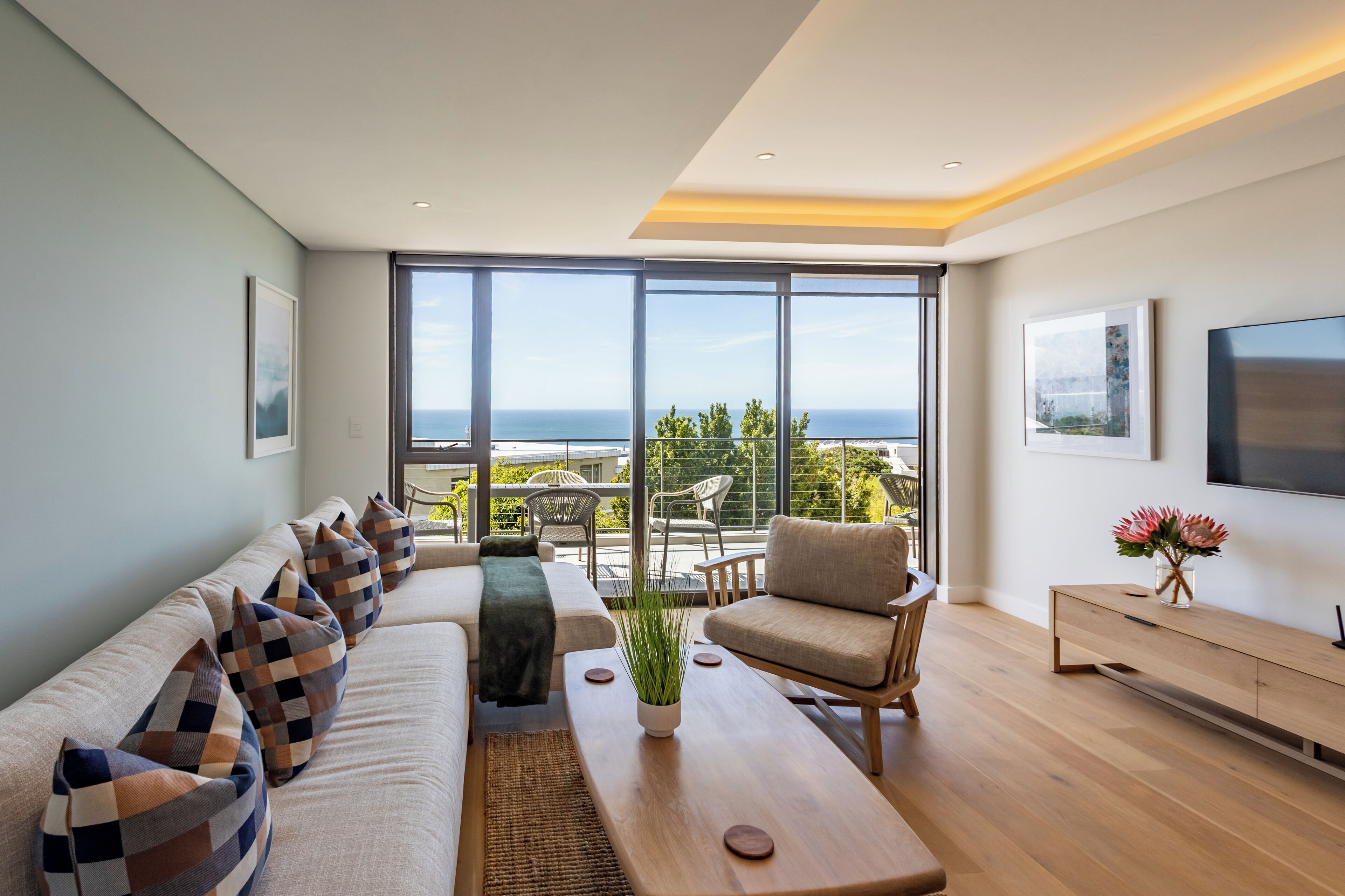 A modern living room with a sofa set, a coffee table, and a TV. There is a balcony with a view of the ocean.