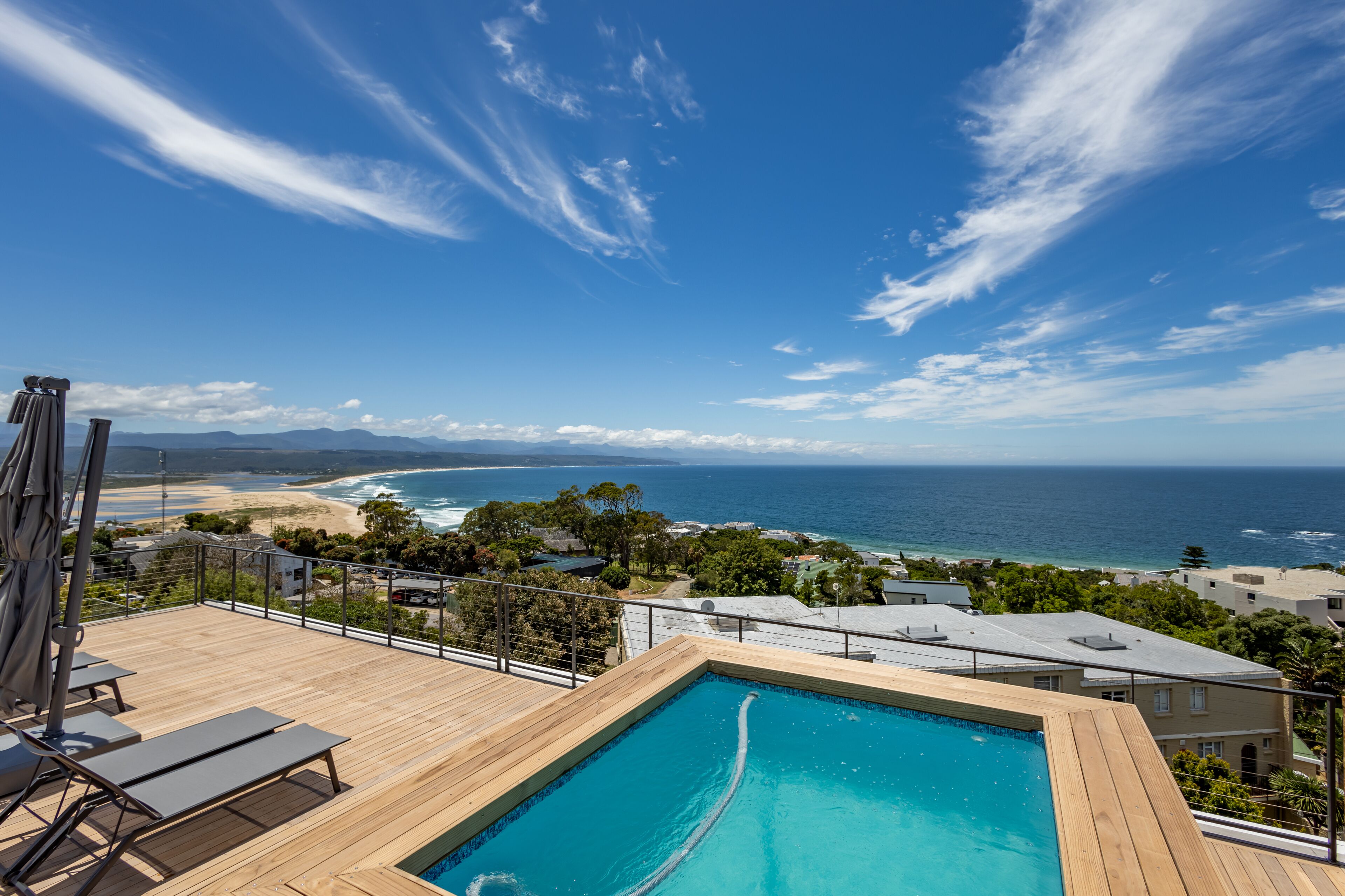 A pool with a view of the ocean and beach, surrounded by greenery and buildings.
