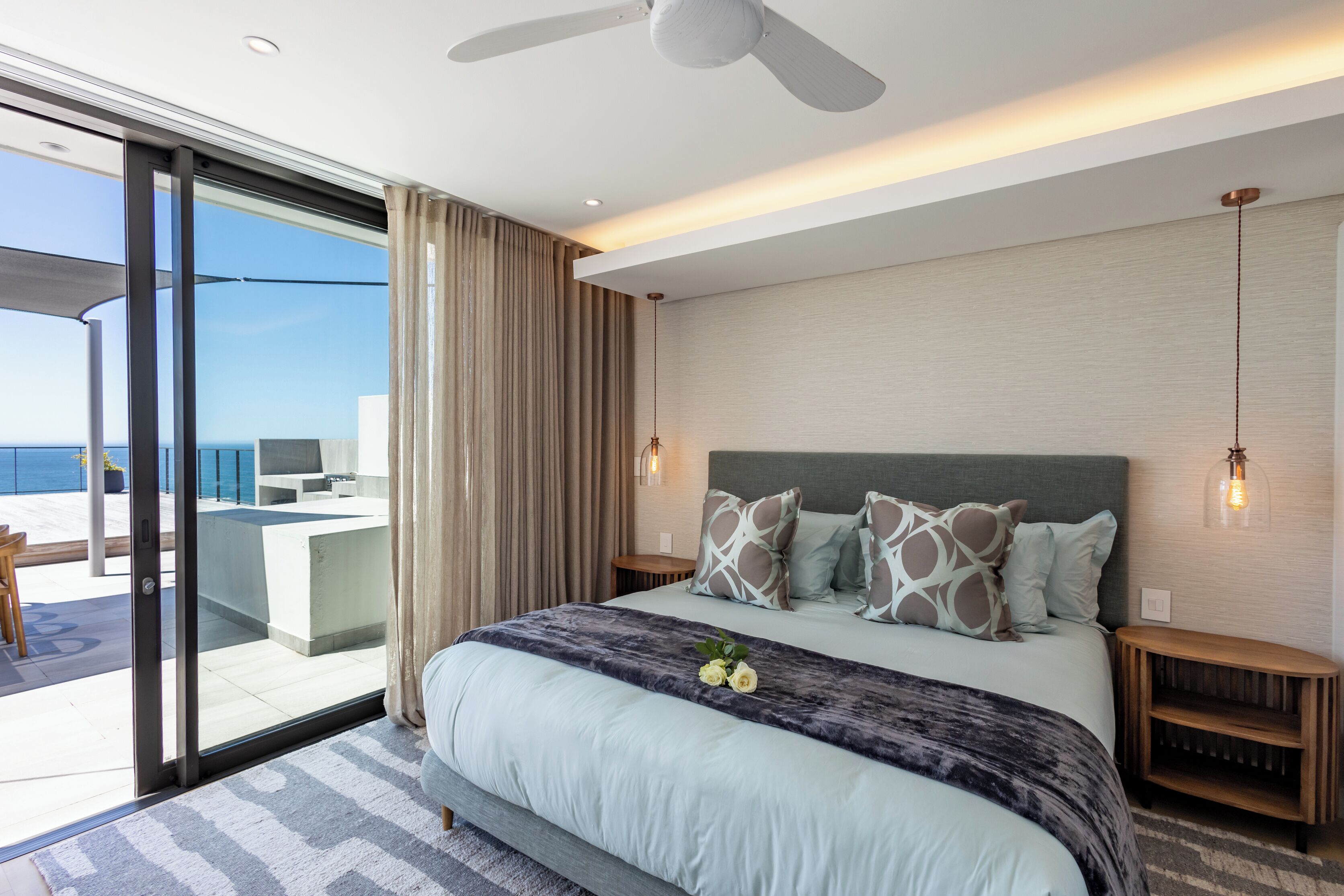 A modern bedroom with a large bed, a ceiling fan, and a view of the ocean through a sliding glass door.