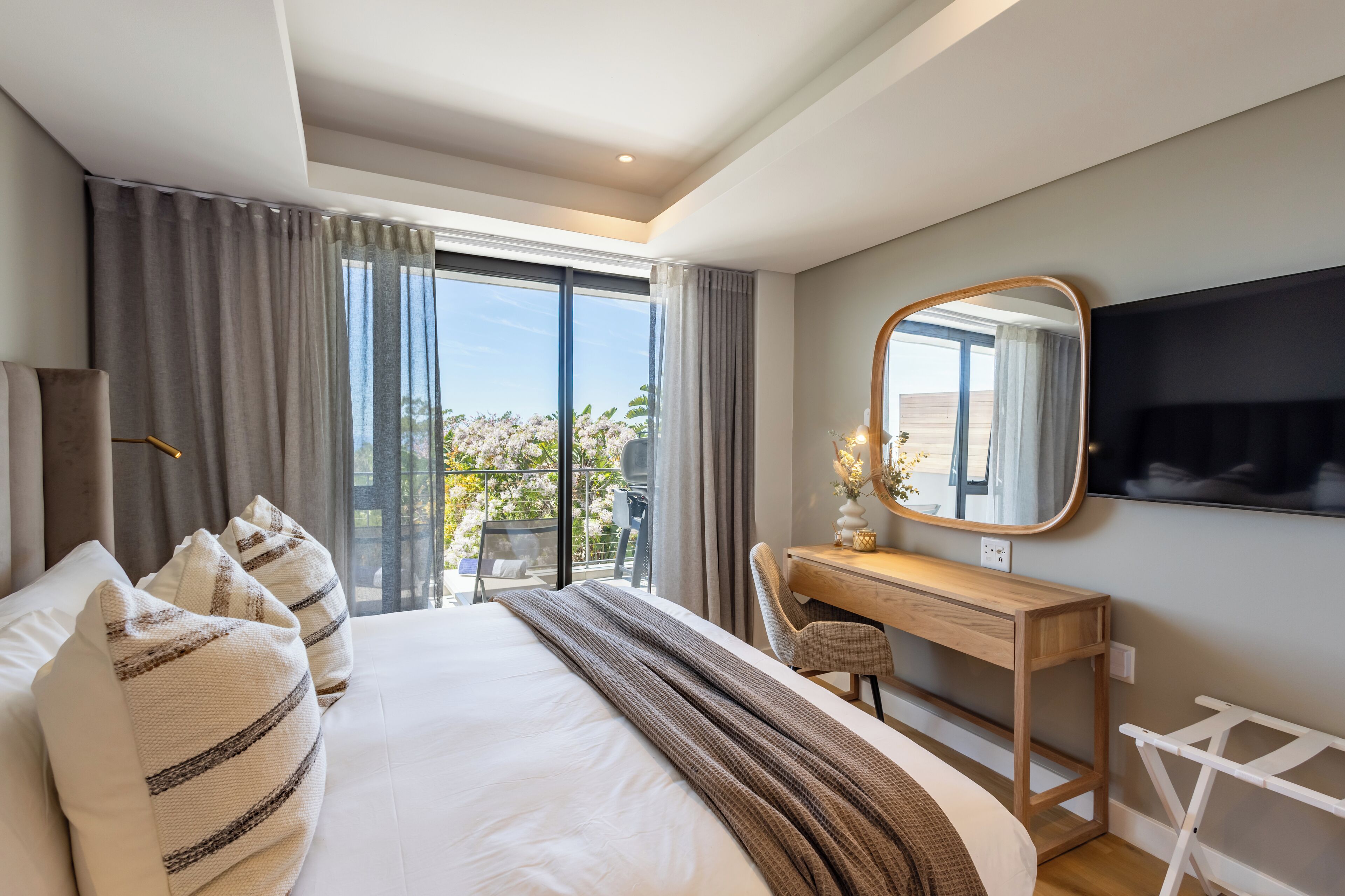 A modern hotel room with a large bed, a wooden desk, a mirror, and a balcony with a view of trees.