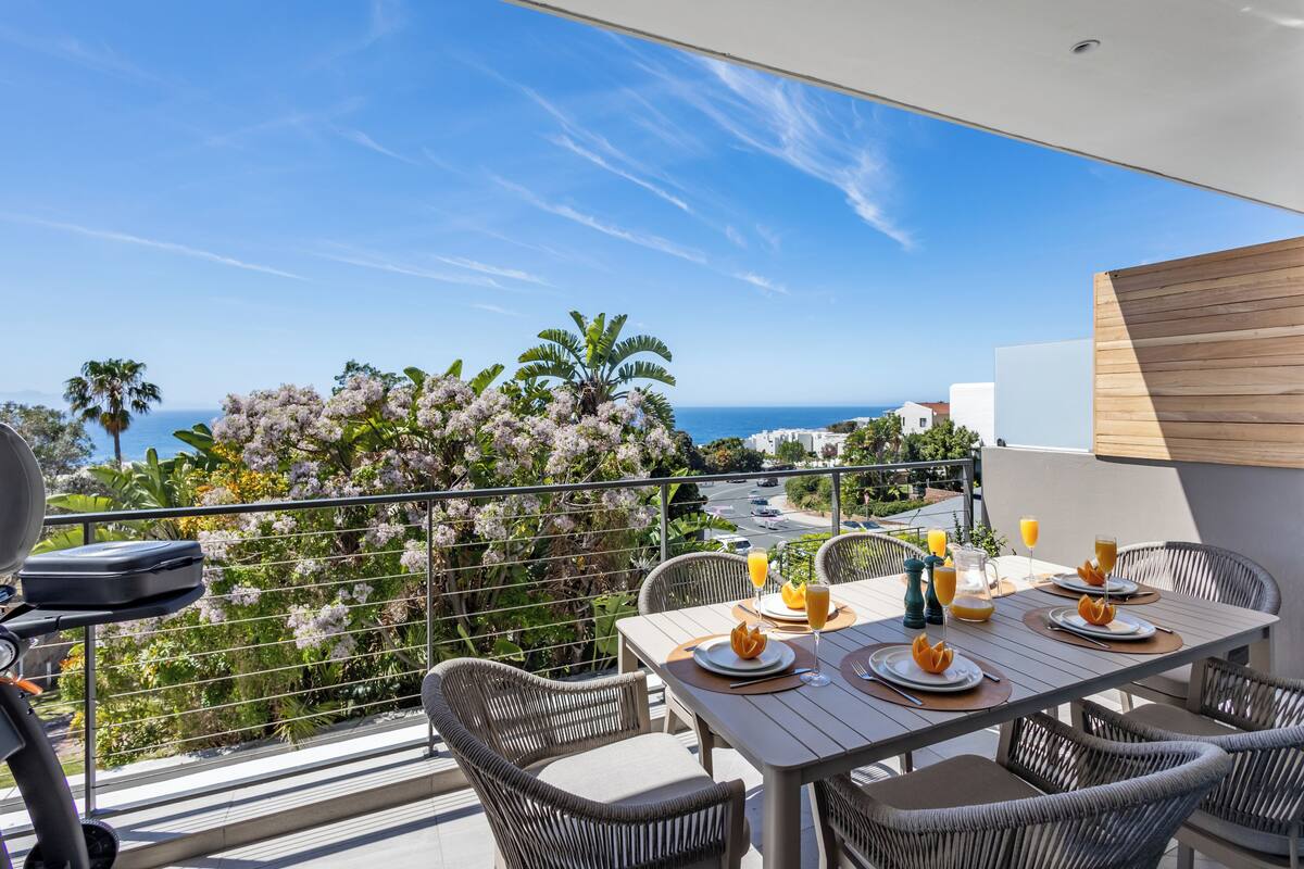 A balcony with wicker furniture, a table set for two with plates, glasses, and a pitcher of orange juice, overlooking a coastal view with palm trees and the sea.