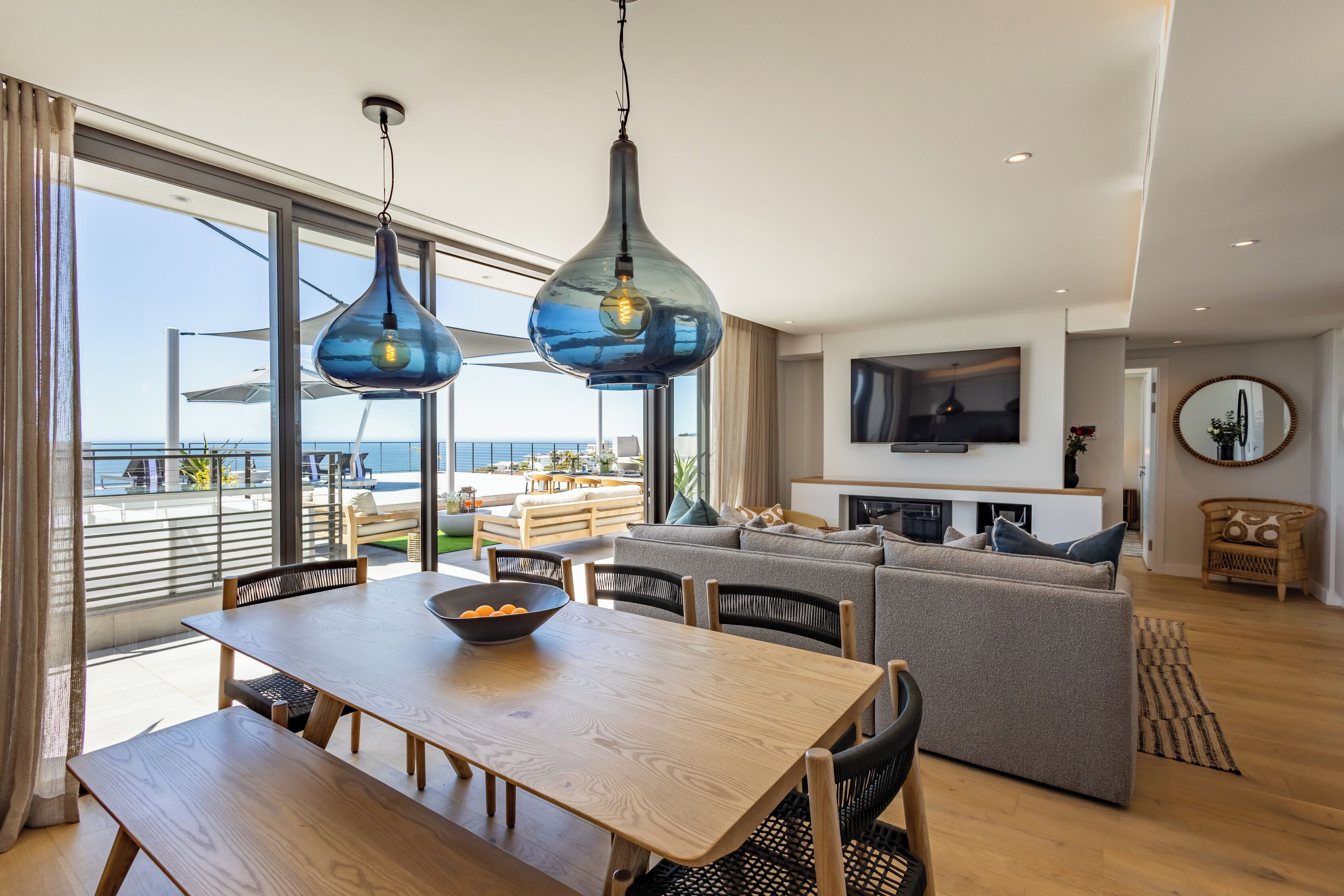 A modern living room with a wooden dining table, grey sofa, and a TV mounted on the wall.