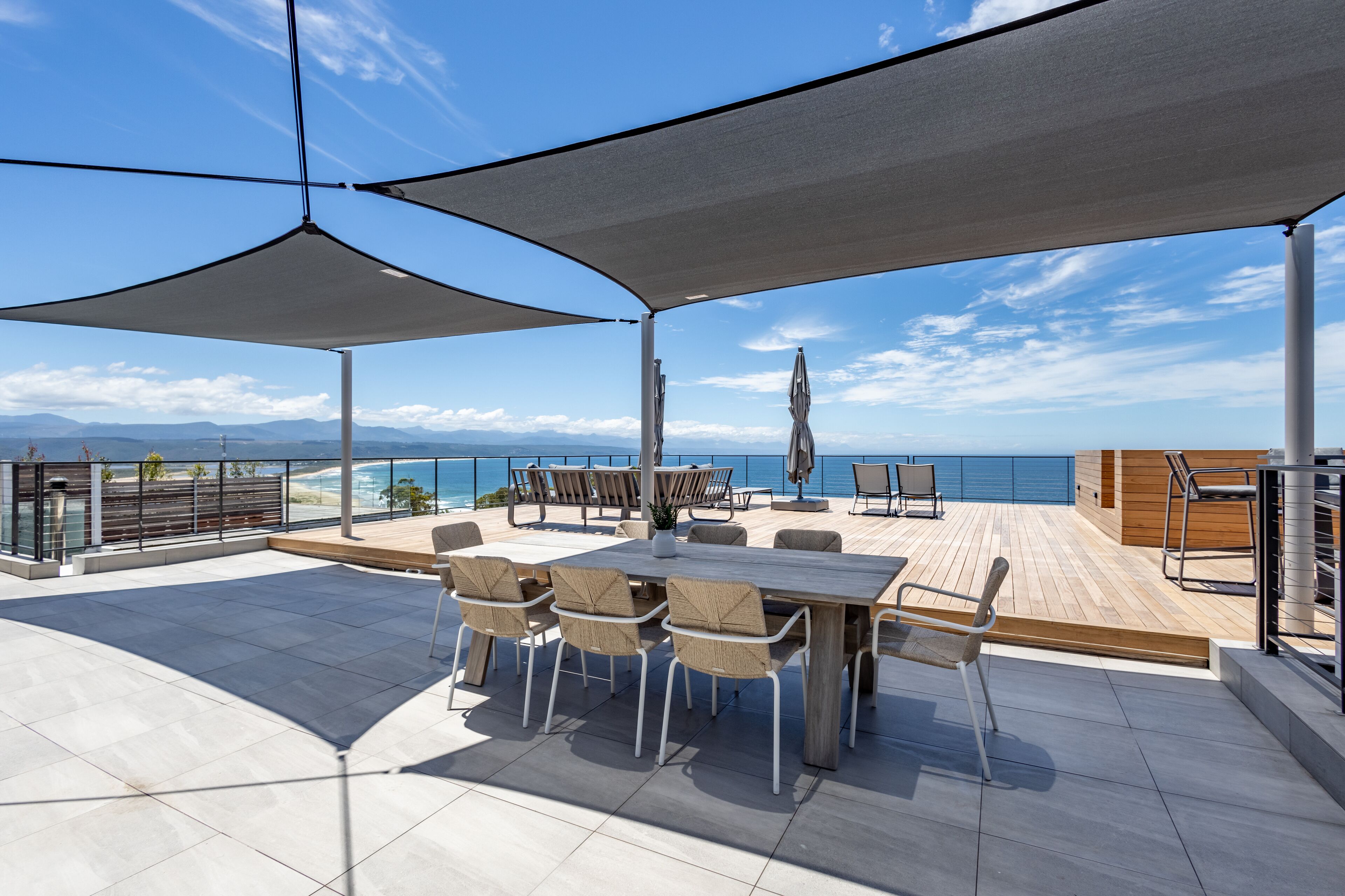 Une terrasse sur le toit, aménagée avec une table à manger, des chaises et des parasols, offrant une vue sur un paysage côtier.