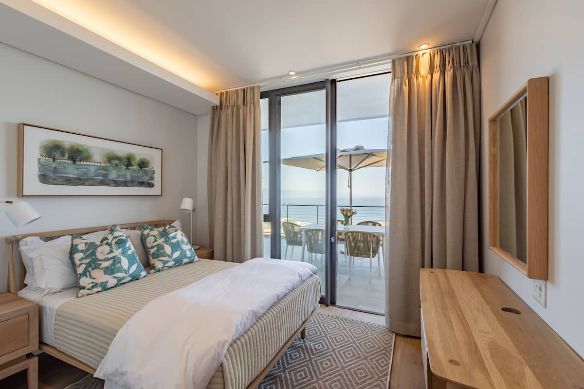 A modern hotel room with a large bed, a wooden nightstand, a desk, and a view of the sea through a sliding glass door.