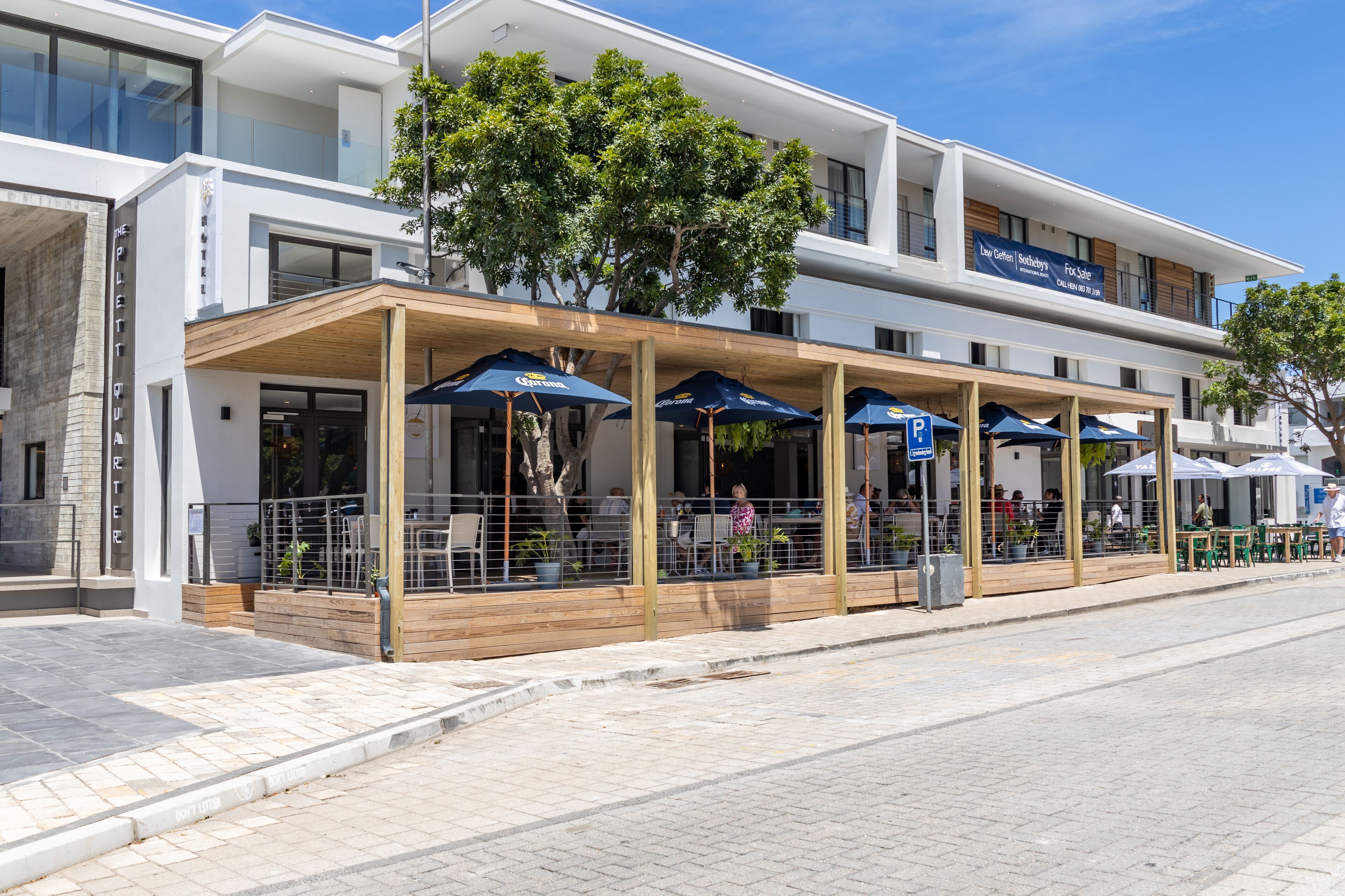 A modern building with a covered outdoor seating area, a sign indicating a sale, and a tree in front.