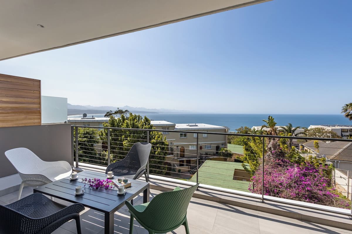 A balcony with a table and chairs, overlooking a coastal view with buildings and a clear sky.