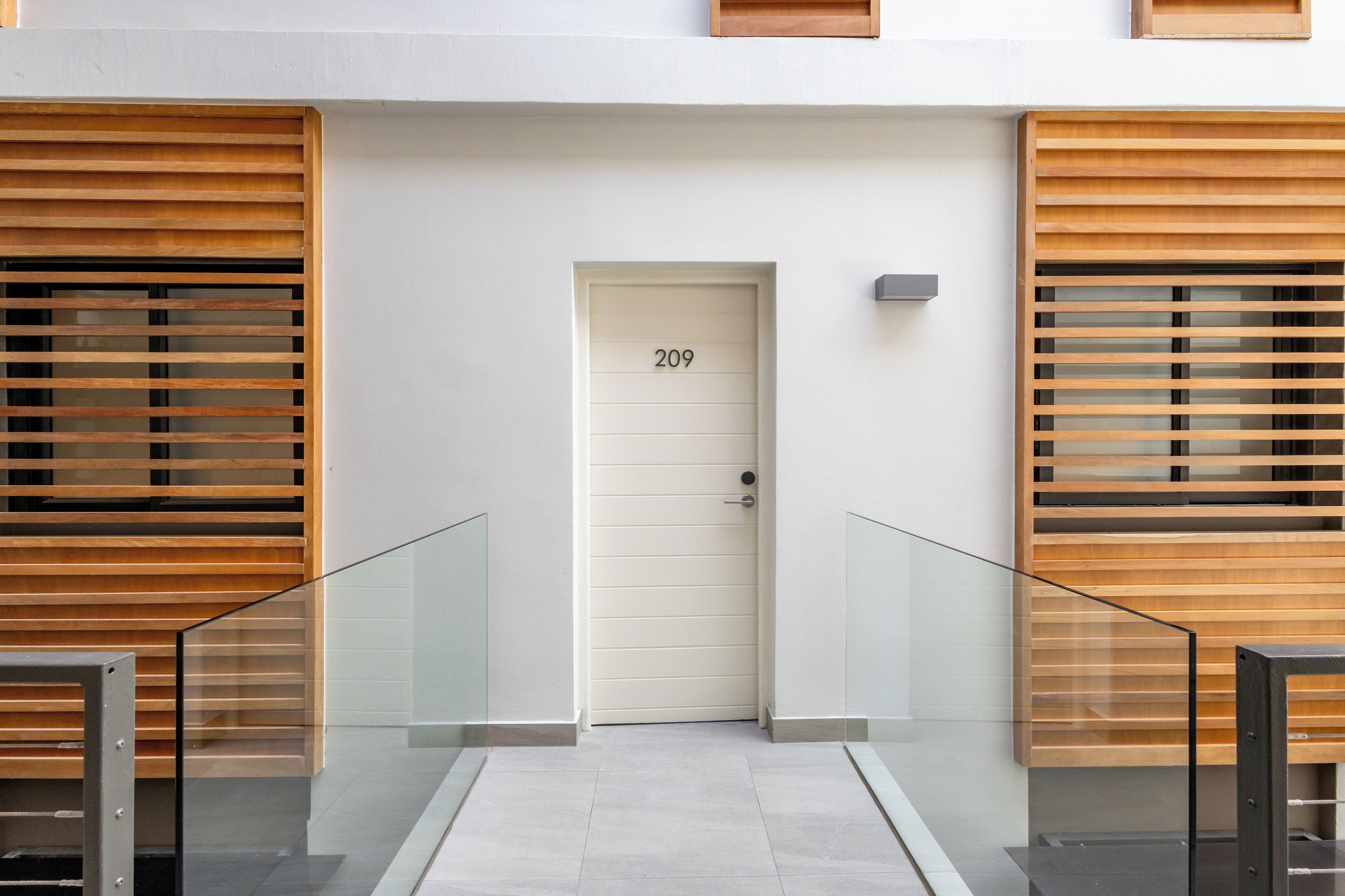 Entrada de un edificio moderno con una puerta blanca marcada “209”, paneles de madera con listones en las paredes y una barandilla de cristal al frente.